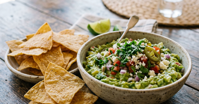 Bowl of guacamole topped with diced tomatoes, onions, cilantro, cheese, and jalapeño next to a bowl of tortilla chips